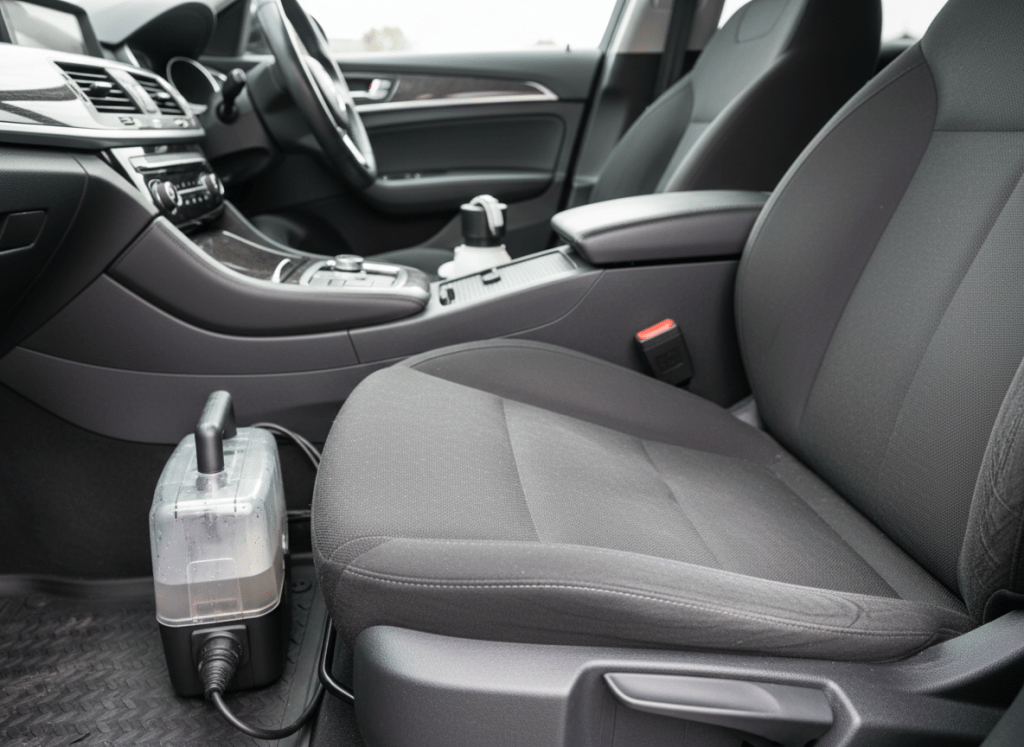 The interior of a vehicle in Belgium just after a professional deep cleaning. A dark grey fabric car seat is shown in close-up, one half perfectly clean and refreshed, the other subtly dusty to demonstrate the before-and-after effect. Fine stitching and fabric texture are rendered with extreme detail. On the floor mat rests a compact professional injection-extraction machine with a transparent upholstery nozzle, still glistening with tiny water droplets. Soft, natural daylight filters through the windshield and side windows, casting controlled highlights on the dashboard and chrome details while leaving the background gently blurred. Photographic realism, shot from the passenger side at an angle that leads the eye along the seat. The mood is meticulous, efficient, and hygienic, underscoring premium vehicle interior cleaning services.